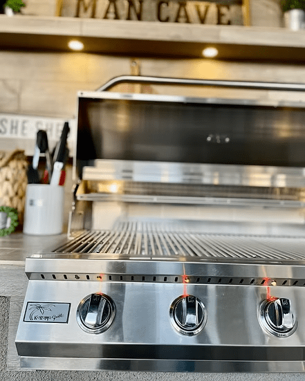 A close-up of the Kokomo Grills Fantasy Kitchen Island 20-Foot by 20-Foot with a 70-Inch TV shows shiny stainless steel, lit burners, open lid, grilling tools nearby, and decor including a MAN CAVE sign on a shelf above the grill.