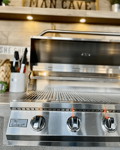 A close-up of the Kokomo Grills Fantasy Kitchen Island 20-Foot by 20-Foot with a 70-Inch TV shows shiny stainless steel, lit burners, open lid, grilling tools nearby, and decor including a MAN CAVE sign on a shelf above the grill.