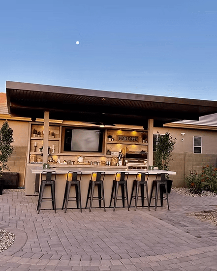 A 20'x20' Kokomo Grills Fantasy Kitchen Island with six black barstools, a 70" TV, decor shelves, and flat roof sits on a paved patio beneath a clear sky and moon.