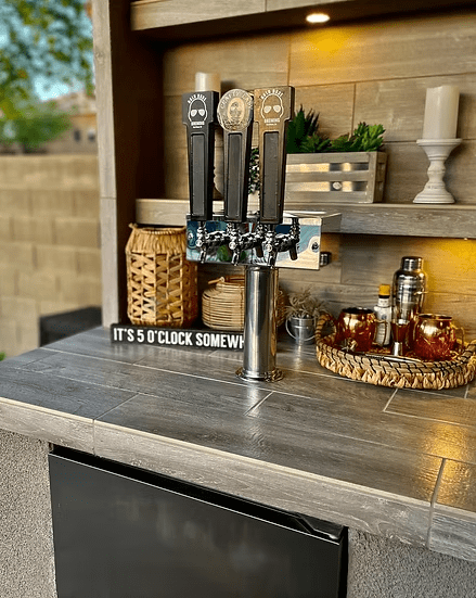 A Kokomo Grills Fantasy Kitchen Island 20’ x 20’ with a 70” TV features three beer taps, an “ITS 5 OCLOCK SOMEWHERE” sign, candles, a cocktail shaker, and copper mugs on a wicker tray atop gray tiled counters—perfect for BBQ gatherings.