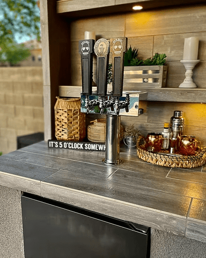 A Kokomo Grills Fantasy Kitchen Island 20’ x 20’ with a 70” TV features three beer taps, an “ITS 5 OCLOCK SOMEWHERE” sign, candles, a cocktail shaker, and copper mugs on a wicker tray atop gray tiled counters—perfect for BBQ gatherings.