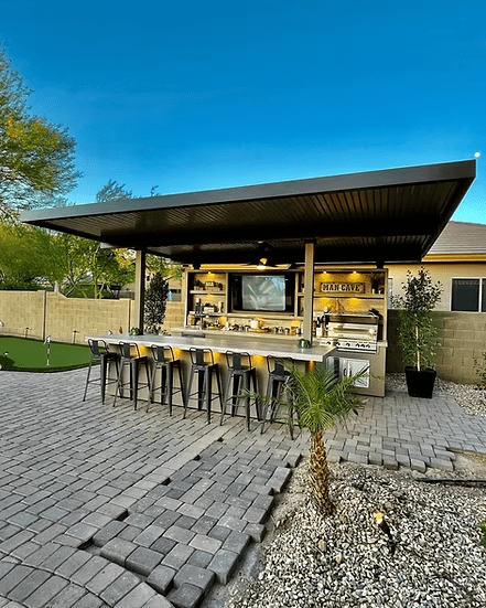 Outdoor bar and kitchen area with the Kokomo Grills Fantasy Kitchen Island 20'x20', 70" TV, barstools, grill, “Man Cave” sign, modern roof, ambient lighting, paving stones, landscaping, and a small palm tree.