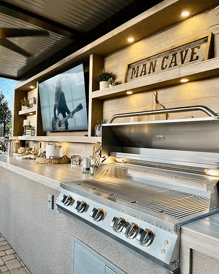 A modern outdoor kitchen with the Kokomo Grills Fantasy Kitchen Island 20’ x 20’, a 70" TV displaying a musician, decorated shelves, plants, a MAN CAVE sign, and recessed lighting beneath a covered patio.