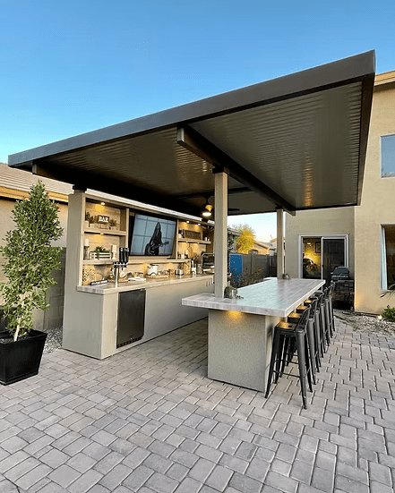 A modern outdoor kitchen showcases the Kokomo Grills Fantasy Kitchen Island 20x20 ft with a 70-inch TV, bar stools, a long countertop, and an overhead roof on a stone patio next to a beige house.