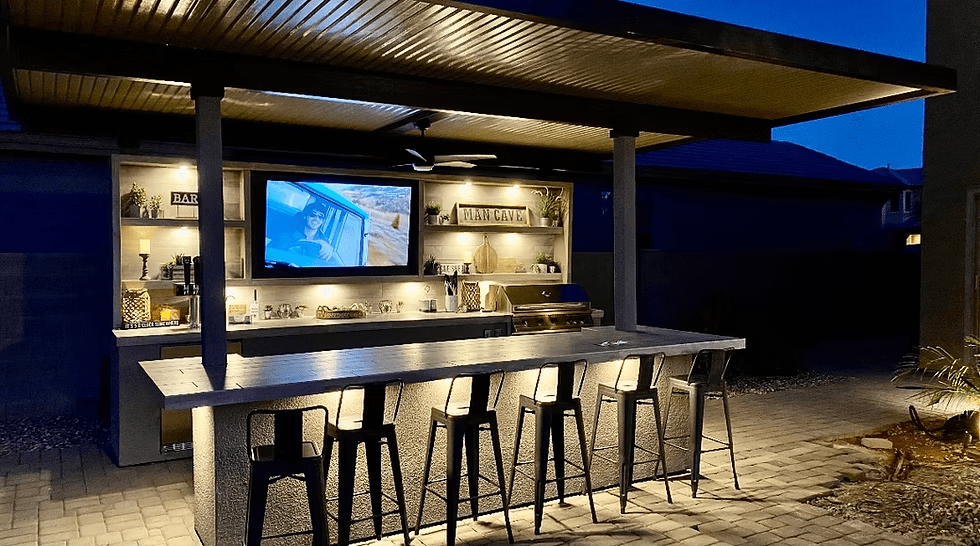 At night, the Kokomo Grills Fantasy Kitchen Island (20' x 20') shines with a built-in grill, glowing countertop, tall metal stools, decor shelves, and a 70-inch wall-mounted TV beneath a wooden pergola.
