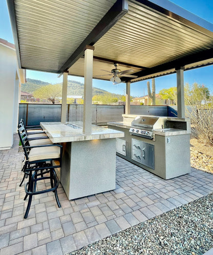 Outdoor kitchen with covered patio, marble countertops, Kokomo Grills Fantasy Kitchen Island 20-Foot by 20-Foot with 70-Inch TV, bar stools, ceiling fan. Stone patio, desert landscaping, and mountain views complete the setting.
