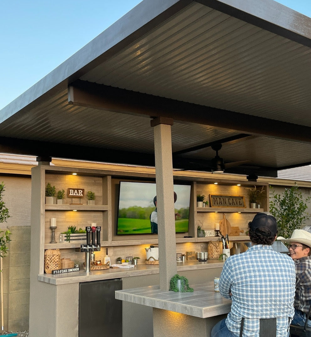 A covered patio outdoor bar features a Kokomo Grills Fantasy Kitchen Island 20-Foot by 20-Foot with a 70-inch TV, decor shelves, beer taps, plants, evening lighting, and two guests in hats at the counter.