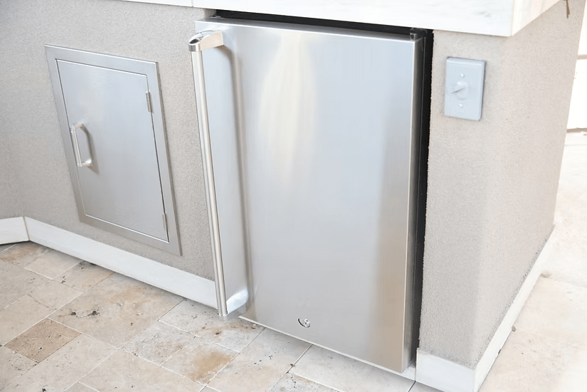 A Kokomo Grills Fridge Door Sleeve is installed into a light-colored outdoor kitchen counter beside a small access panel on a tiled floor.