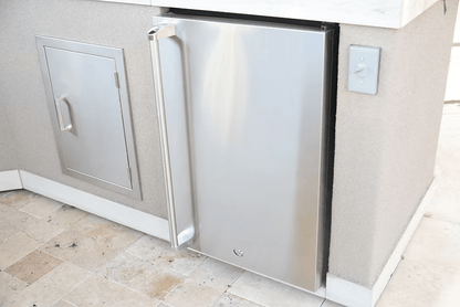 A Kokomo Grills Fridge Door Sleeve is installed into a light-colored outdoor kitchen counter beside a small access panel on a tiled floor.