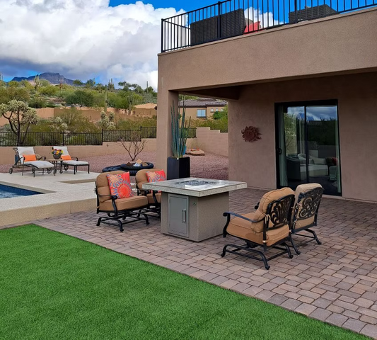 Patio with cushioned chairs and a Kokomo Grills Jamaica Fire Table with fire-glass by artificial grass on pavers. In the background: a pool, lounge chairs, desert landscaping, and mountains beneath a partly cloudy sky.