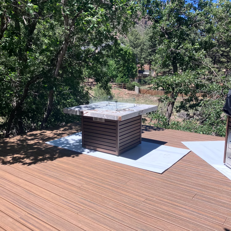 A Kokomo Grills Jamaica Fire Table with Fire-Glass and a glass wind guard sits on a sunny wooden deck surrounded by trees, with white mats underneath for an inviting setting.