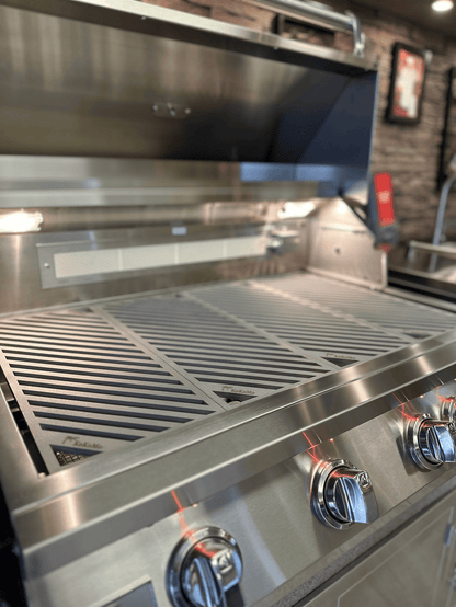 A close-up of a stainless steel Kokomo Grills gas grill with the lid open, featuring LuxCut Laser-Cut Culinary Grates for 3, 4, or 5 burner Classic and Pro models and four control knobs, set in a modern kitchen or showroom.