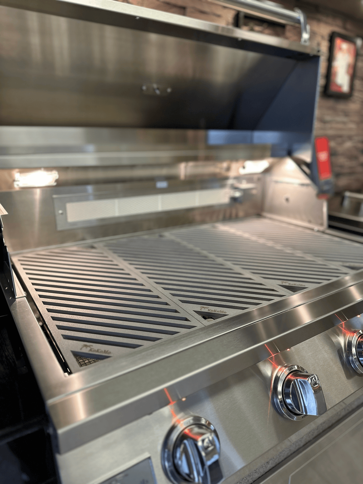 A close-up of a Kokomo Grills gas grill with the lid open, showcasing Kokomo Grills LuxCut Laser-Cut Culinary Grates and three front control knobs, set in a modern kitchen or patio.