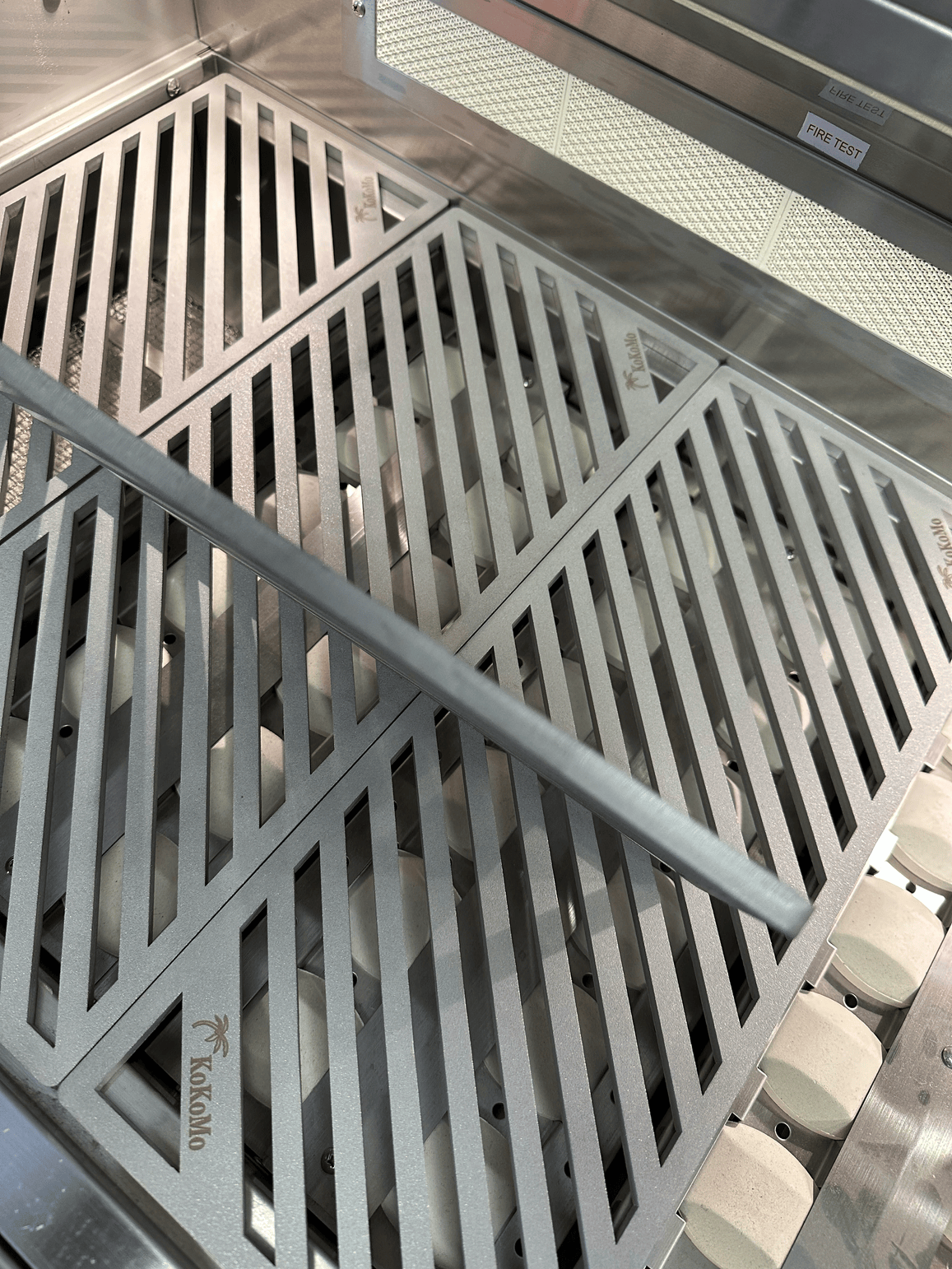 Close-up of Kokomo Grills LuxCut Laser-Cut Culinary Grates for 3, 4, 5 Burner Classic and Pro Grills, showing diagonal slots, a metal bar on the 304 stainless steel surface, and distinctive Kokomo branding etched on the grates.