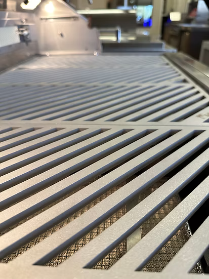 Close-up of Kokomo Grills LuxCut Laser-Cut Culinary Grates for 3, 4, and 5 burner Classic and Pro grills. The modern diagonal-slatted metal grate stands out amid a blurred commercial kitchen background with industrial equipment.