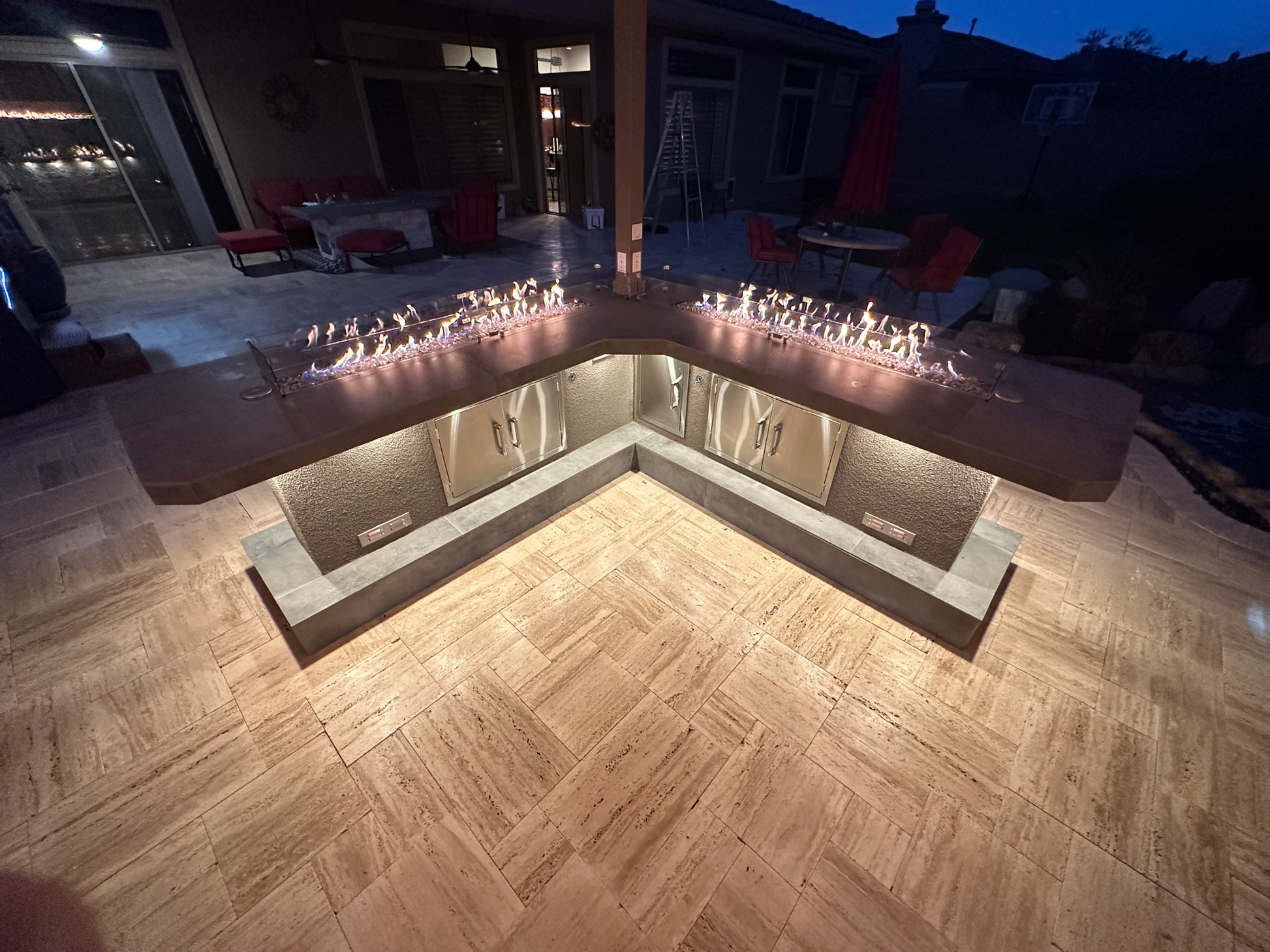 At dusk, the Kokomo Grills Luxury Outdoor Kitchen BBQ Grill Island features an L-shaped stone counter, built-in cabinets, warm lighting, a gas fire feature, and patio furniture for a functional and inviting outdoor space.
