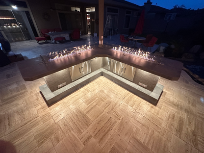 At dusk, the Kokomo Grills Luxury Outdoor Kitchen BBQ Grill Island features an L-shaped stone counter, built-in cabinets, warm lighting, a gas fire feature, and patio furniture for a functional and inviting outdoor space.