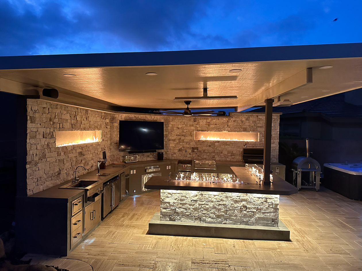 A modern outdoor kitchen by Kokomo Grills features the Kokomo Grills Luxury Outdoor Kitchen BBQ Grill Island, stone walls, built-in appliances, under-counter lighting, a central island, linear fire features on the wall, and ceiling fans below a flat roof.