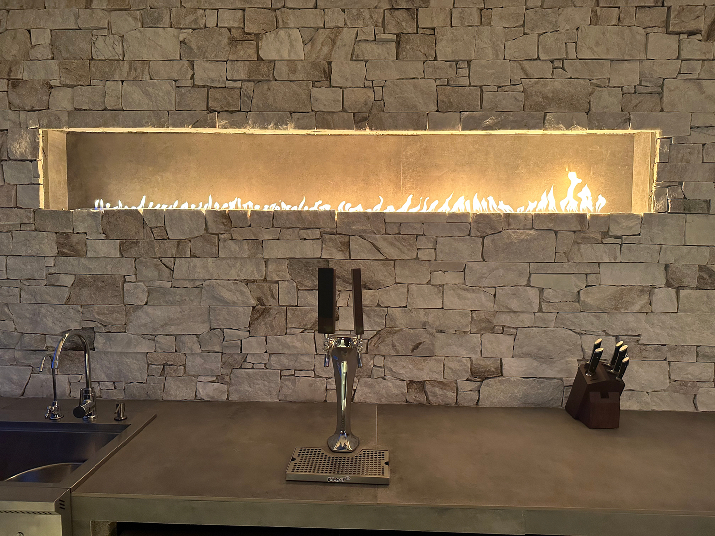 The Kokomo Grills Luxury Outdoor Kitchen BBQ Grill Island features a stone wall, horizontal gas fireplace, countertop with sink and beer tap, wooden knife block, and all essentials for a premium grilling experience.