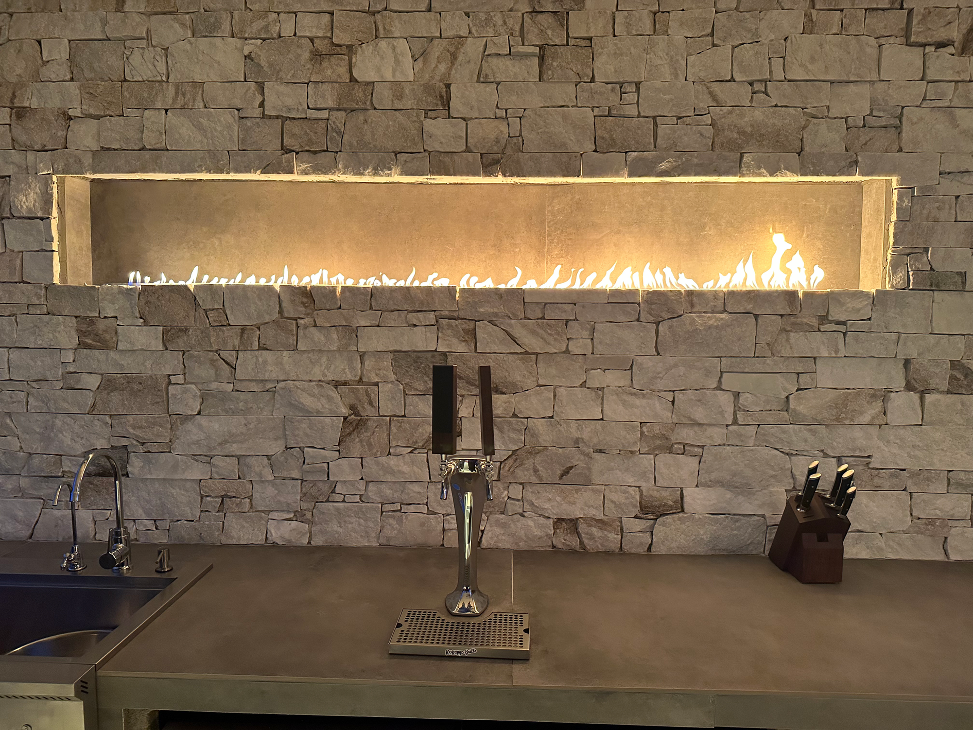 The Kokomo Grills Luxury Outdoor Kitchen BBQ Grill Island features a stone wall, horizontal gas fireplace, countertop with sink and beer tap, wooden knife block, and all essentials for a premium grilling experience.