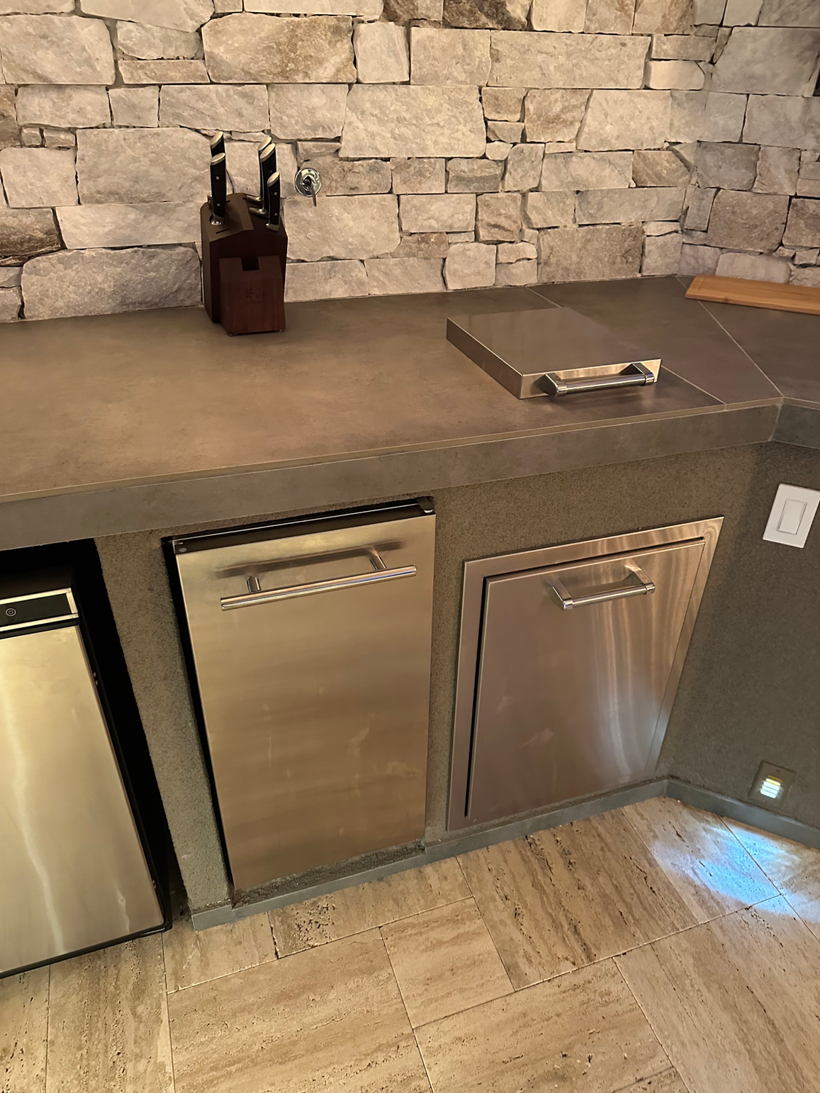 A modern kitchen countertop with knife block, metal tray, and stone backsplash sits above stainless steel dishwasher and cabinets—ideal for pairing with the Kokomo Grills Luxury Outdoor Kitchen BBQ Grill Island by Kokomo Grills.