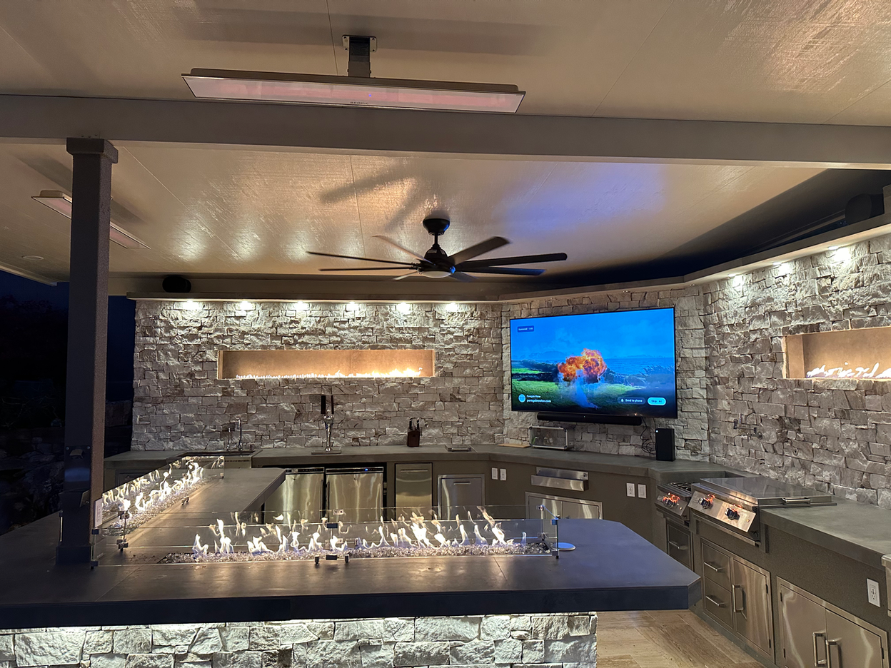 The Kokomo Grills Luxury Outdoor Kitchen BBQ Grill Island features stone walls, a built-in grill, sink, countertops, linear fire feature along the bar, ambient lighting, ceiling fan, and a wall-mounted TV playing a movie.