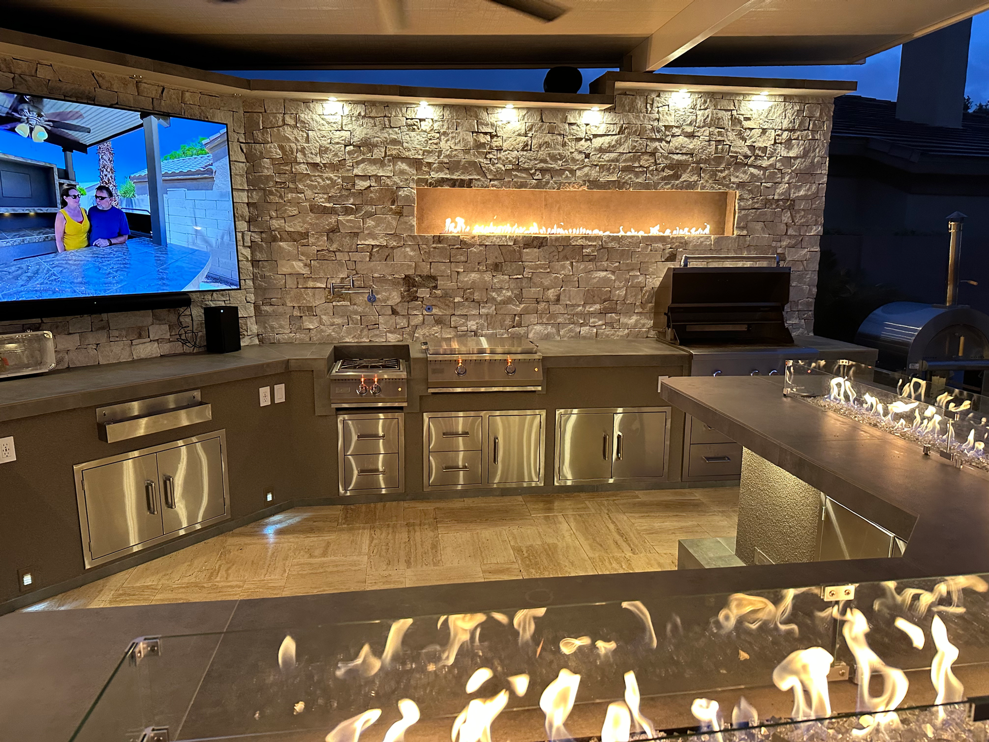 The Kokomo Grills Luxury Outdoor Kitchen BBQ Grill Island features stainless steel appliances, a stone wall with a gas fireplace, TV, and glass fire features—perfect for modern outdoor entertaining at dusk.