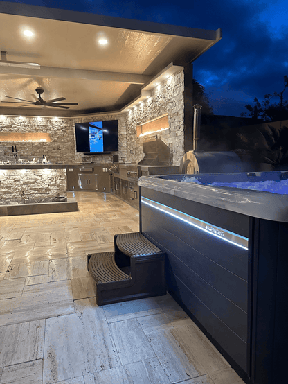 Outdoor kitchen and lounge area with stone walls, TV, ceiling fan, modern lighting, and a Kokomo Grills Luxury Outdoor Kitchen BBQ Grill Island. A hot tub with steps and blue LED lights sits on the tiled patio at dusk.