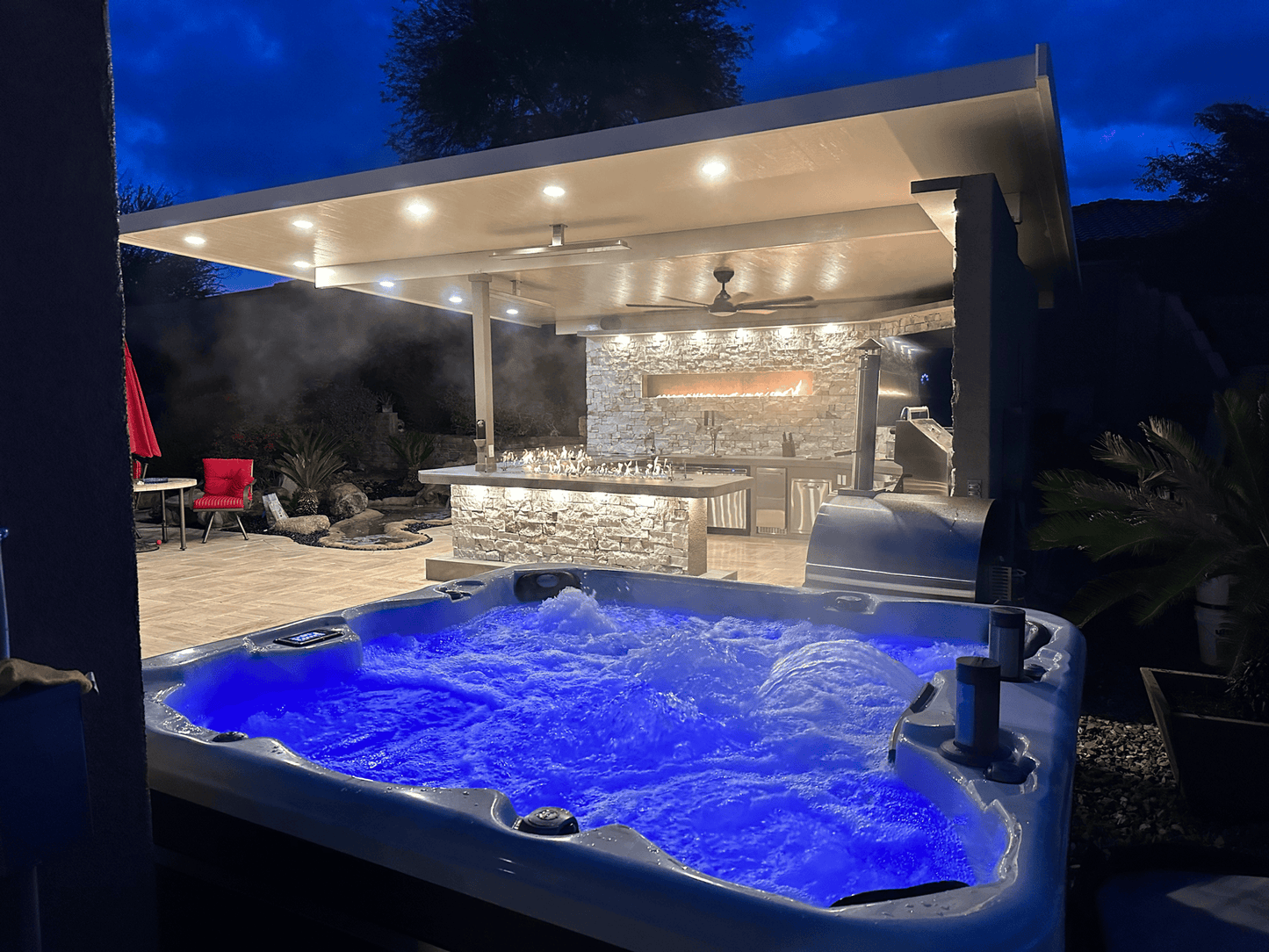A hot tub with blue LED bubbles sits in front, while a Kokomo Grills Luxury Outdoor Kitchen BBQ Grill Island by Kokomo Grills, featuring stone accents and soft lighting, creates an inviting retreat beneath the evening sky.