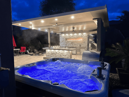 A hot tub with blue LED bubbles sits in front, while a Kokomo Grills Luxury Outdoor Kitchen BBQ Grill Island by Kokomo Grills, featuring stone accents and soft lighting, creates an inviting retreat beneath the evening sky.