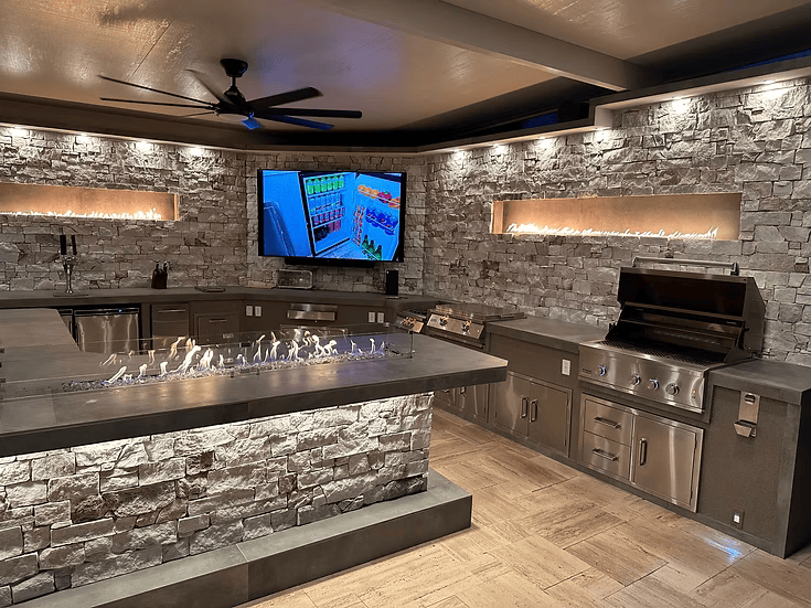 The Kokomo Grills Luxury Outdoor Kitchen BBQ Grill Island by Kokomo Grills features stone walls, countertops, sink, bar area, large TV, overhead fan, and decorative linear fire elements—enhanced with soft lighting for a modern outdoor experience.