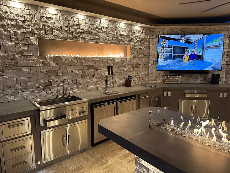 The Kokomo Grills Luxury Outdoor Kitchen BBQ Grill Island features a stone accent wall, built-in gas fireplace, stainless steel appliances, under-counter sink, mounted TV with pool view, and an island with glass fire feature in the foreground.