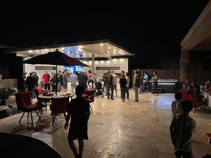 A lively group socializes at night on a well-lit patio featuring the Kokomo Grills Luxury Outdoor Kitchen BBQ Grill Island, with tables and chairs. Some guests stand and chat, others sit, and kids are present—creating a relaxed atmosphere.