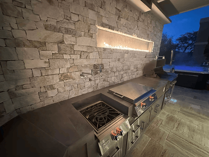 The Kokomo Grills Luxury Outdoor Kitchen BBQ Grill Island by Kokomo Grills features stainless steel countertops, glowing burner flames, drawers, sleek stone wall accents, and overhead lighting—ideal for luxury grill enthusiasts enjoying twilight views.