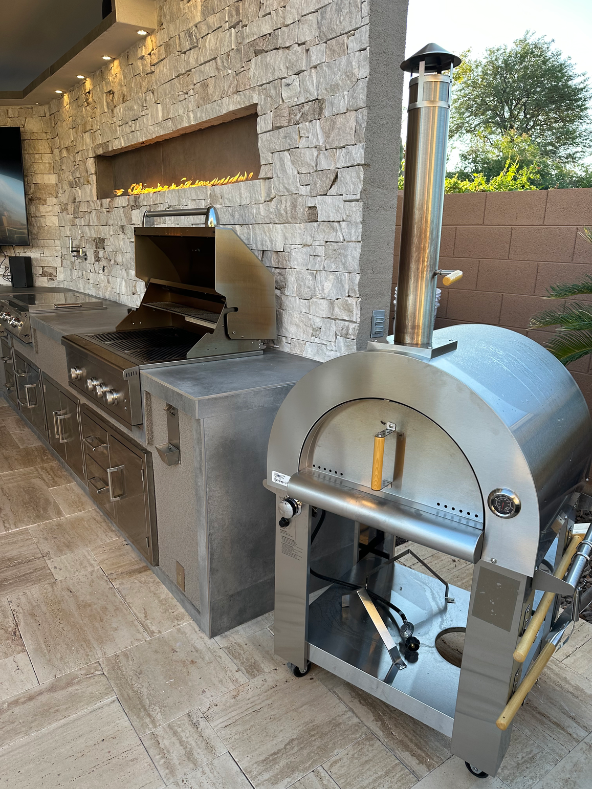 A modern outdoor kitchen features a Kokomo Grills Luxury Outdoor Kitchen BBQ Grill Island, stainless steel appliances, a pizza oven with chimney, stone backsplash, built-in cabinets, and a wall-mounted gas fireplace. Natural light and trees are visible outside.