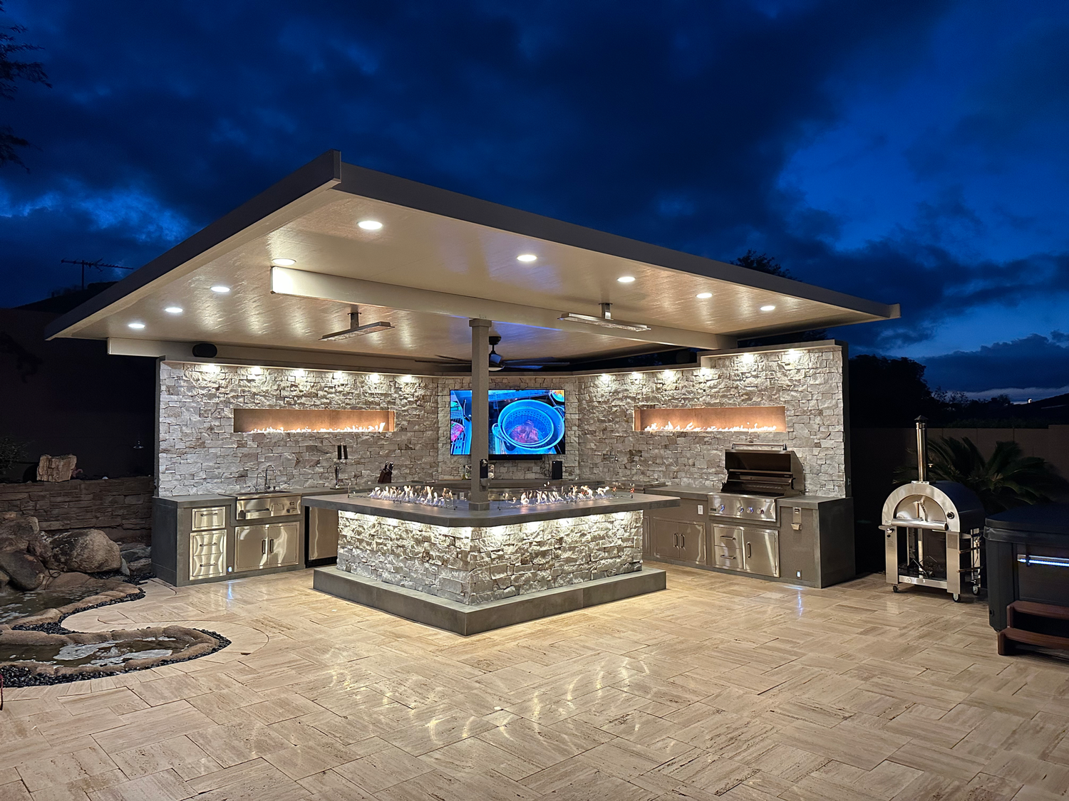 The Kokomo Grills Luxury Outdoor Kitchen BBQ Grill Island by Kokomo Grills features stone walls, stainless steel appliances, under-cabinet lighting, and a TV above the counter—perfect for evening gatherings under a flat roof at dusk.
