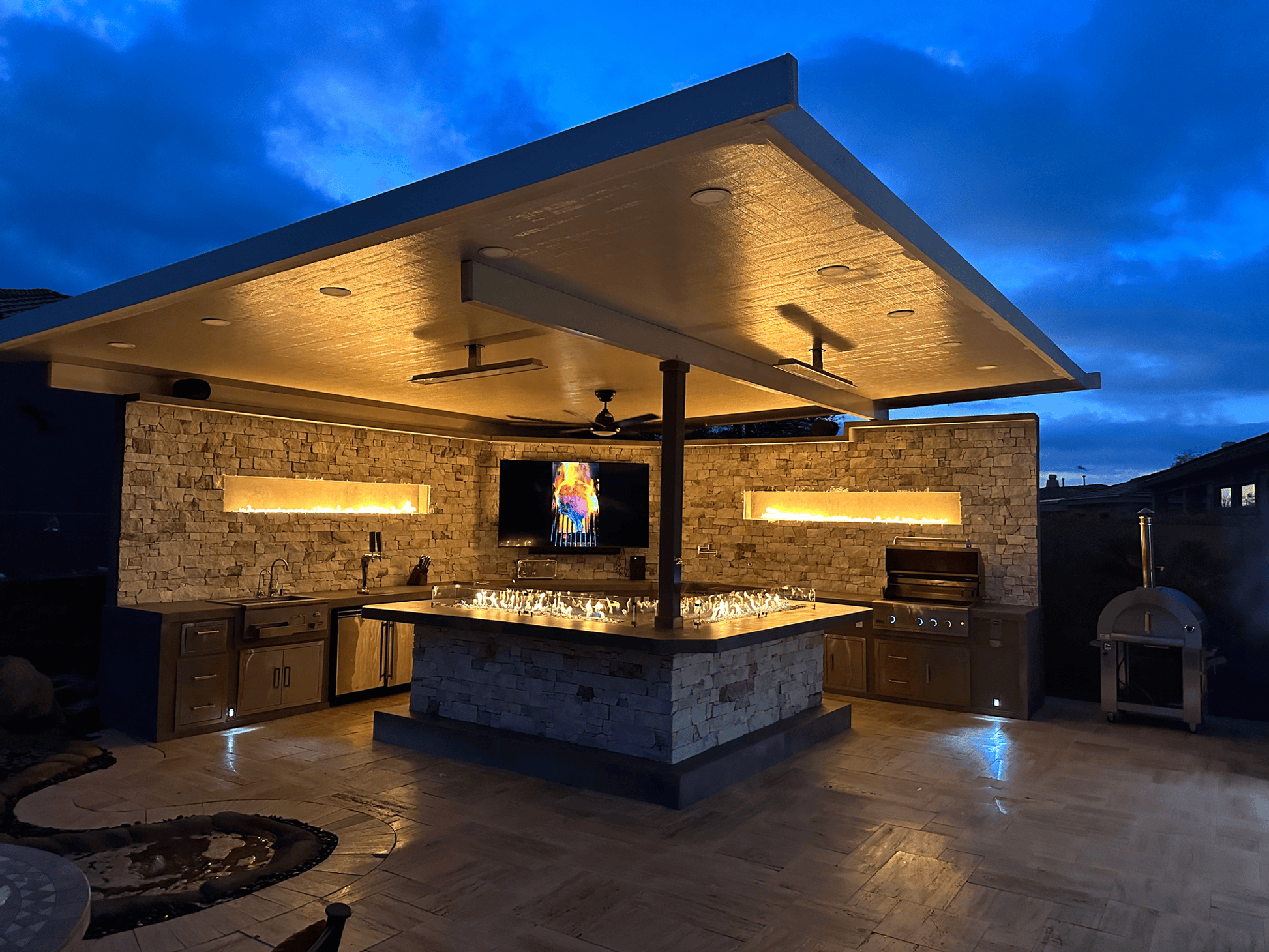 At dusk, the Kokomo Grills Luxury Outdoor Kitchen BBQ Grill Island features stone walls, built-in cabinets, a luxury grill, pizza oven, and TV above an illuminated countertop. Warm lights highlight the grill island under a flat roof for a cozy vibe.