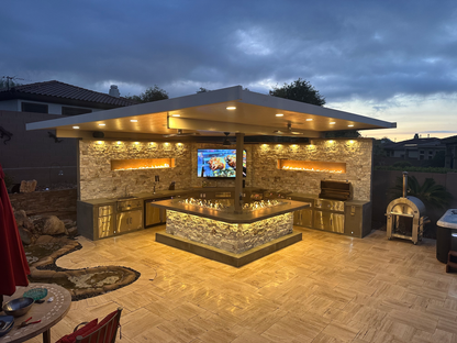 A modern outdoor kitchen by Kokomo Grills features the Kokomo Grills Luxury Outdoor Kitchen BBQ Grill Island, stone counters, and pizza oven under a flat roof. Warm lighting and a TV above the counter create a cozy dusk atmosphere.