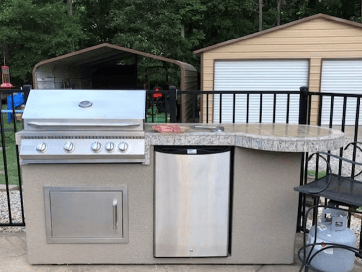 Outdoor kitchen features a Kokomo Grills Maui 7′6” BBQ Island with a 33” round bar, built-in BBQ, LED lights, and stainless steel details. The fenced area also includes storage sheds and background trees.