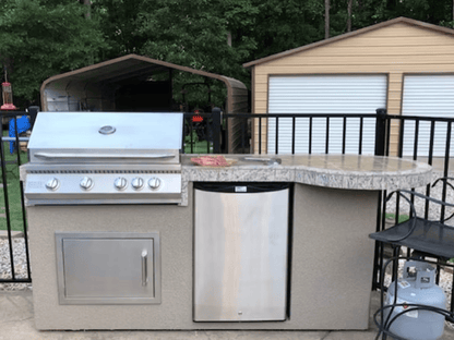 Outdoor kitchen features a Kokomo Grills Maui 7′6” BBQ Island with a 33” round bar, built-in BBQ, LED lights, and stainless steel details. The fenced area also includes storage sheds and background trees.