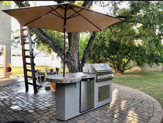 The Kokomo Grills Maui 7′6” BBQ Island with 33” Round Bar, LED lights, and built-in BBQ by Kokomo Grills is showcased under a patio umbrella on a brick patio, with trees and lawn creating a scenic backdrop.