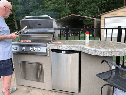 An older man in a blue shirt and shorts grills meat on a Kokomo Grills Maui 7′6” BBQ Island with 33” round bar, LED lights, and built-in BBQ in an outdoor kitchen with refrigerator and counter; trees and sheds are in the background.