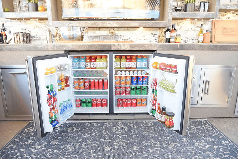Two open mini fridges under the Kokomo Grills Outdoor Kitchen Montego Model BBQ Island and Grill are filled with drinks, condiments, fruits, and vegetables. Shelves with kitchen items are visible above.