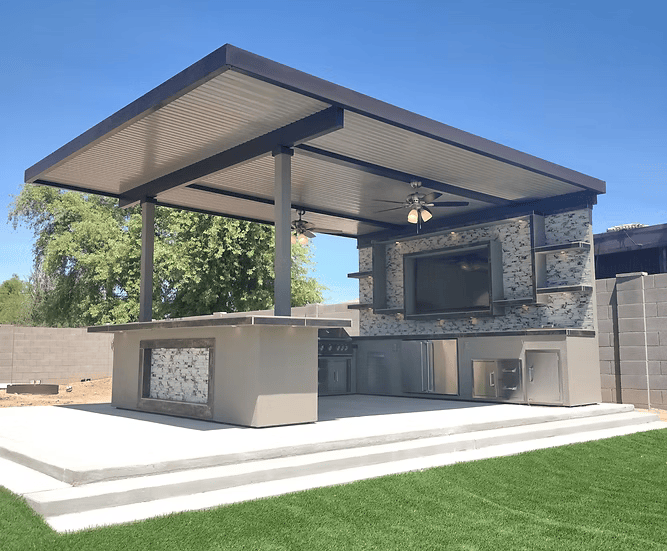 Modern outdoor kitchen with a flat roof, ceiling fans, TV, and the Kokomo Grills Outdoor Kitchen Montego Model BBQ Island and Grill by Kokomo Grills on a concrete patio surrounded by grass, trees, and a gray block fence.