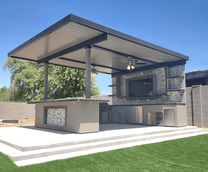 Modern outdoor kitchen with a flat roof, ceiling fans, TV, and the Kokomo Grills Outdoor Kitchen Montego Model BBQ Island and Grill by Kokomo Grills on a concrete patio surrounded by grass, trees, and a gray block fence.