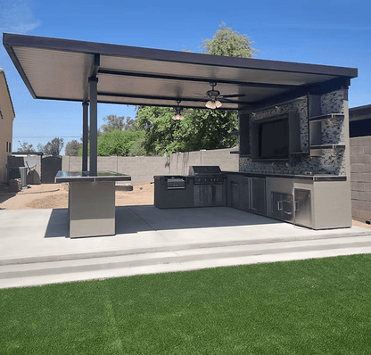 Enjoy a modern outdoor kitchen with the Kokomo Grills Outdoor Kitchen Montego Model BBQ Island and Grill, sink, and TV under a covered patio with ceiling fans. The space features a concrete patio, artificial grass, block wall fence, trees, and blue sky.