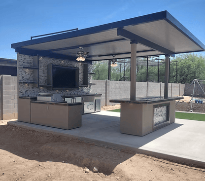 The Kokomo Grills Montego Model BBQ Island and Grill creates a stylish outdoor kitchen with TV, counter space, ceiling fans, and bar area under a metal roof on a concrete patio with block wall and playground in view.