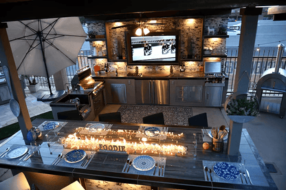 Modern outdoor kitchen and dining area at dusk, showcasing a long table set with blue and white plates, overhead lights, stainless steel appliances, TV, decorative accents, and the Kokomo Grills Outdoor Kitchen Montego Model BBQ Island and Grill as the focal point.