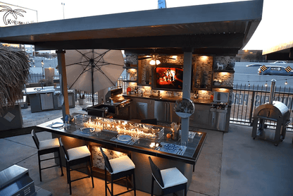 Modern and stylish, the Kokomo Grills Outdoor Kitchen Montego Model BBQ Island features bar seating, a countertop fire feature, covered TV area, umbrella, premium appliances, evening lighting, and a fenced backdrop for a stunning setup.