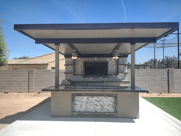 Outdoor bar and kitchen with a Kokomo Grills Outdoor Kitchen Montego Model BBQ Island and Grill, metal roof, stone accents, TV, ceiling fans, and black countertops on a concrete patio with a block wall and houses in the background.